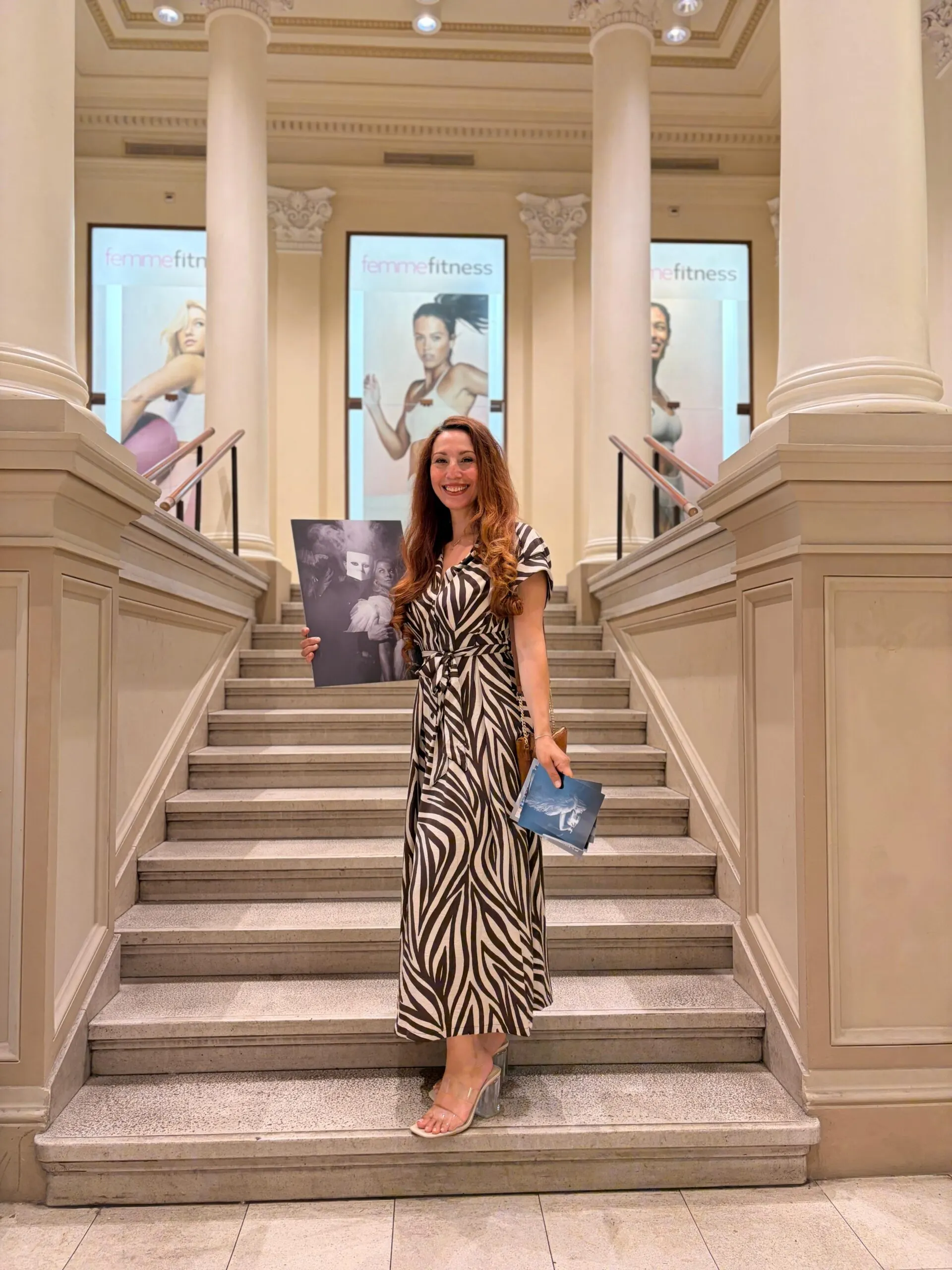 Frau im Zebramuster-Kleid hält Schwarzweiß-Fotodruck auf einer Freitreppe mit Femme-Fitness-Plakaten