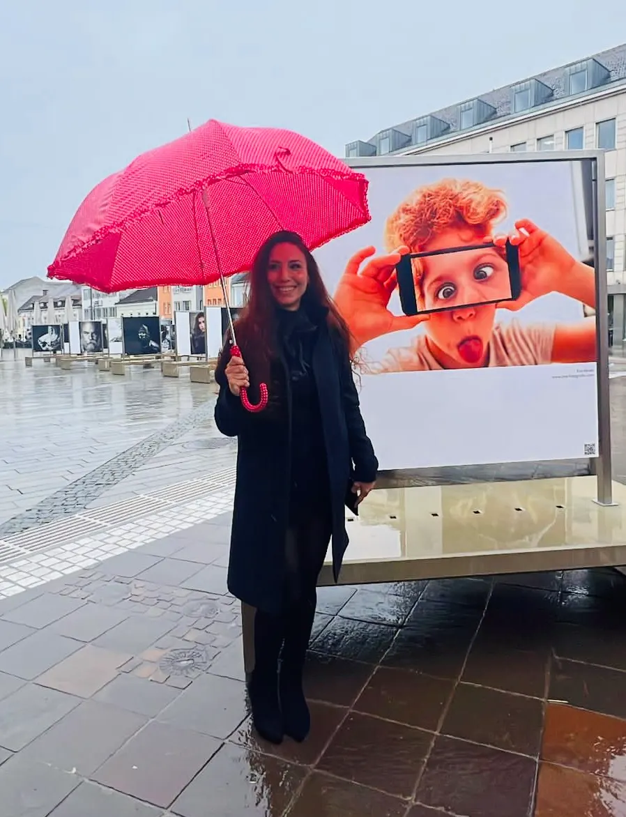 Frau mit rotem Schirm vor Open-Air-Ausstellungstafel mit Kinderfoto auf regennassem Stadtplatz