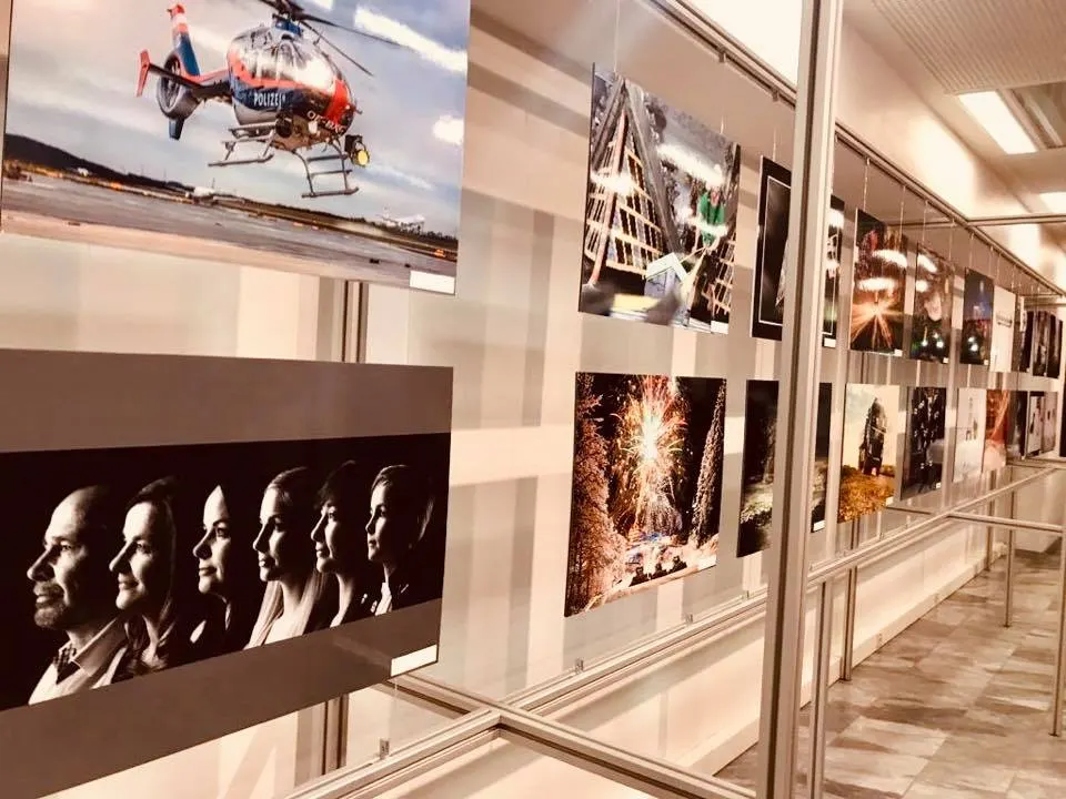 Ausstellungsgang mit hängenden Fototafeln, darunter Polizeihubschrauber, Feuerwerk und Schwarzweiß-Porträtreihe