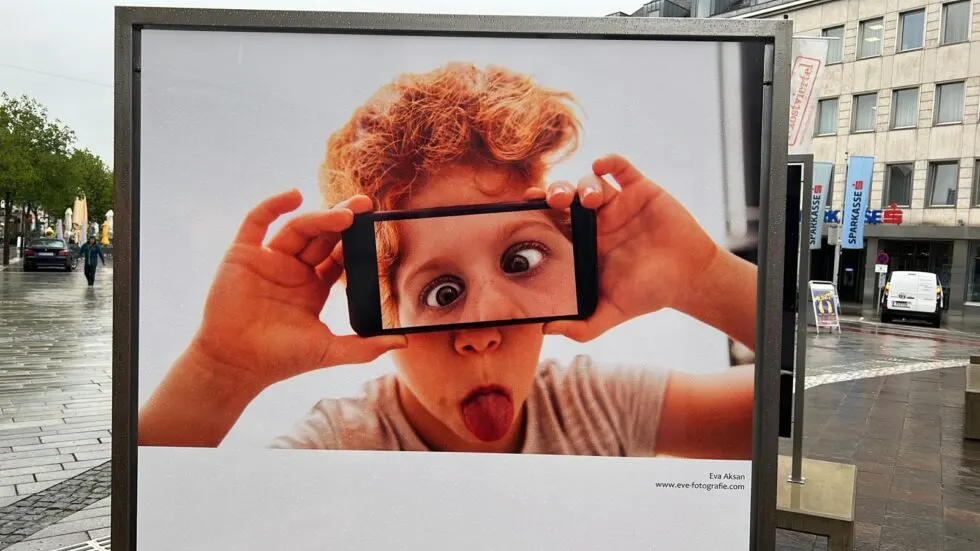 Ausstellungstafel auf einem Platz zeigt Foto eines rothaarigen Buben, der ein Smartphone vor die Augen hält, Bildtext Eva Aksan