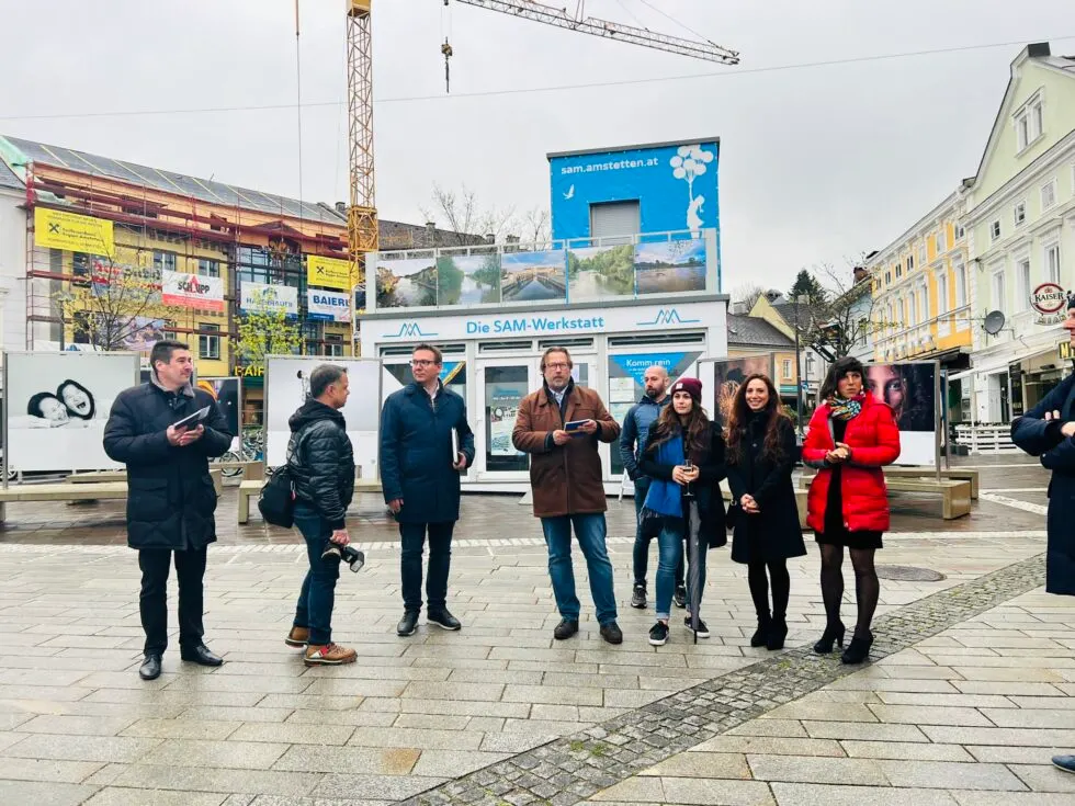 Vernissagegäste zwischen Ausstellungstafeln mit Fotografien auf einem Stadtplatz, dahinter Baukran und Gebäude