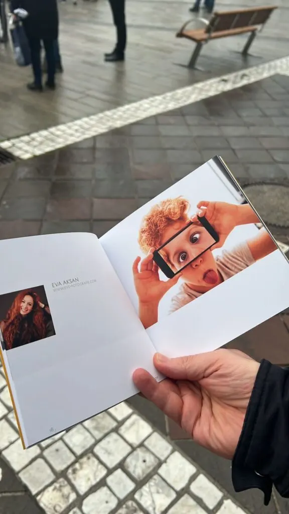 Hand hält aufgeschlagenen Katalog mit Seite von Eva Aksan, daneben Foto eines Buben mit Smartphone