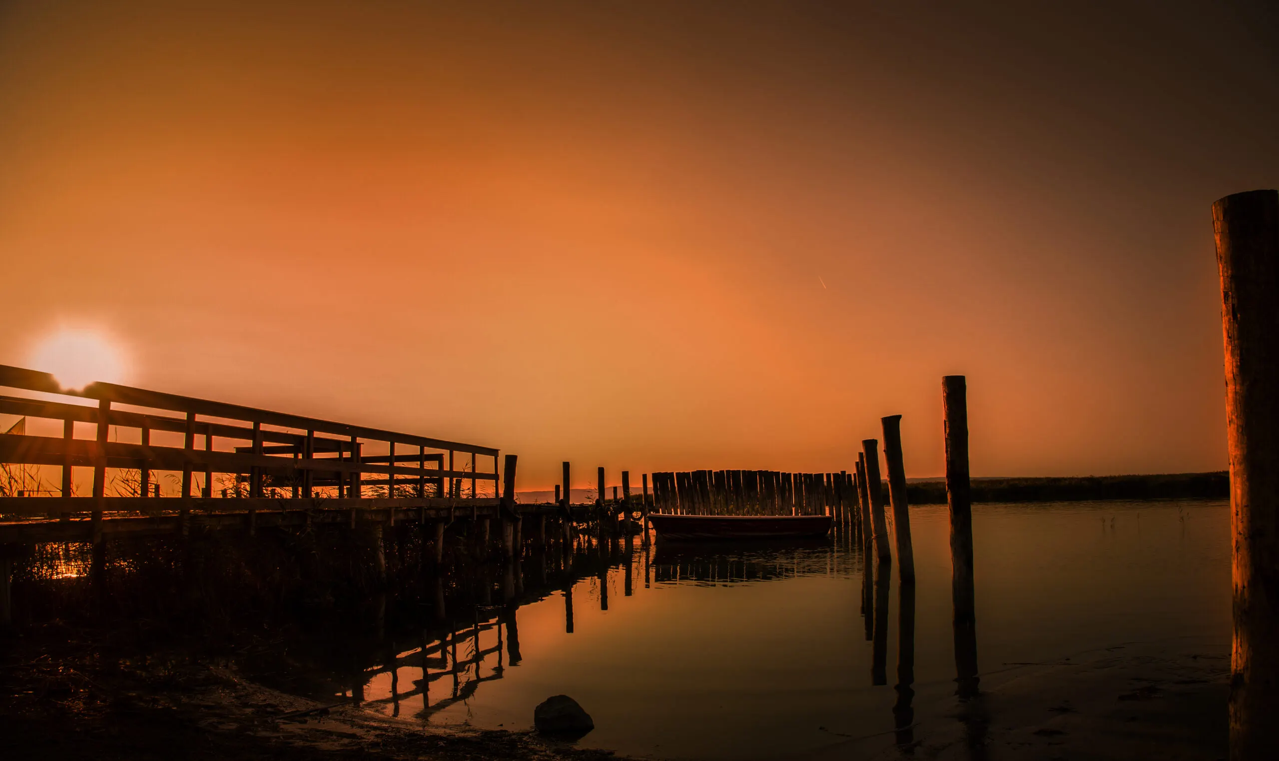 Sonnenuntergang am See mit Holzsteg, Ruderboot und alten Pfählen im stillen Wasser