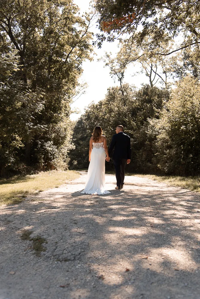 Hand in Hand gehen Angel und Christoph einen sonnigen Kiesweg unter hohen Bäumen entlang, von hinten aufgenommen