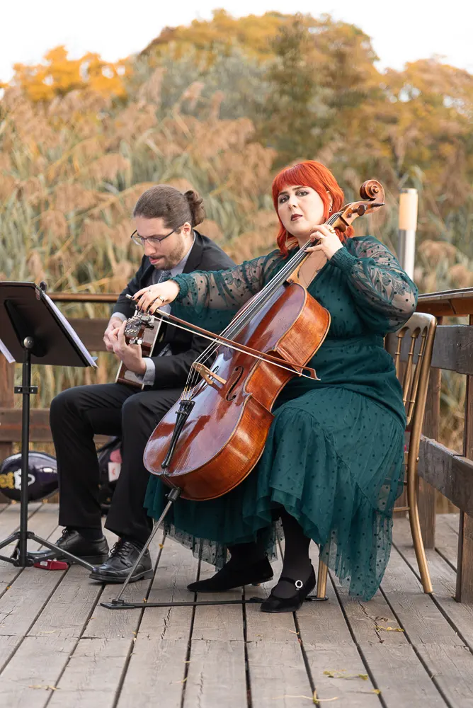 Cellistin im grünen Tüllkleid und Gitarrist begleiten die Trauung auf dem Holzdeck vor herbstlichem Schilf.