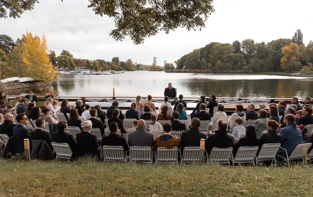 Trauung am Ufer: Gäste auf weißen Stühlen blicken zum Wasser, dahinter Boote, Herbstbäume und Hochhäuser am Horizont.