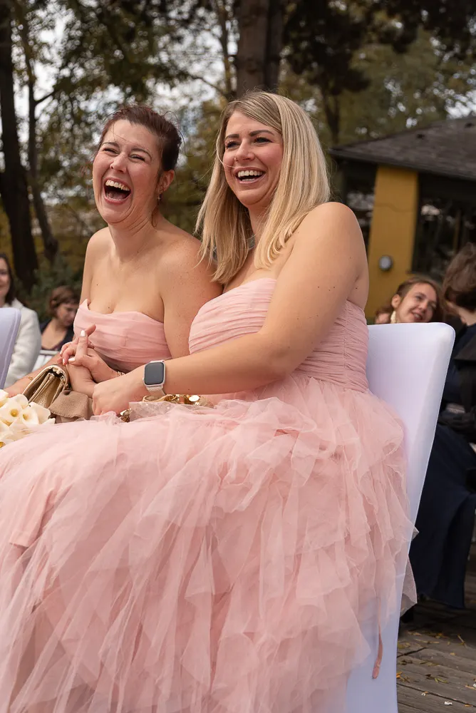 Die Freundinnen in Rosa lachen befreit während der Zeremonie, Calla-Strauß und goldene Tasche im Schoß.