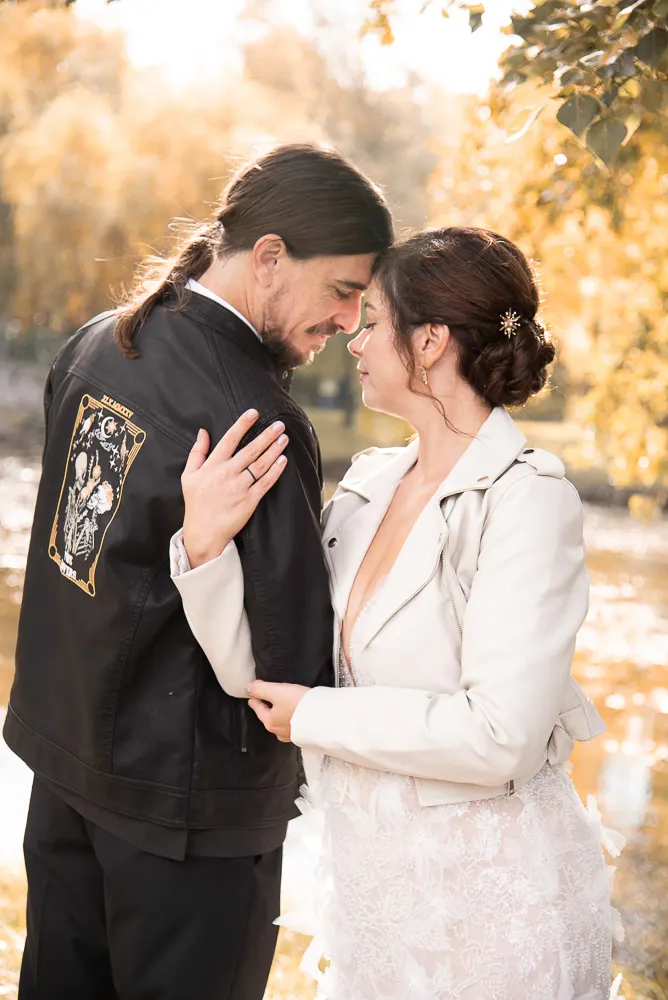 Lederjacken über der Hochzeitskleidung: er in Schwarz mit Golddruck, Angi in Creme, Stirn an Stirn am Ufer.