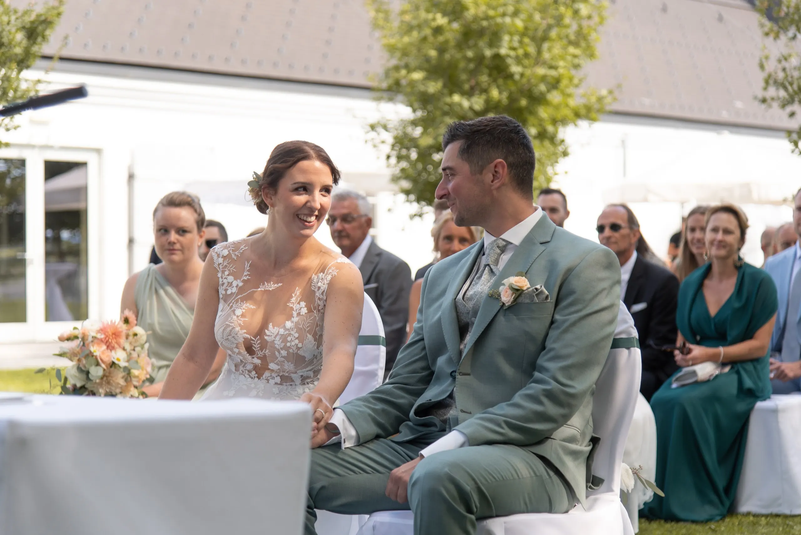 Anja und Martin sitzen bei der freien Trauung im Freien, sie lacht ihn an, er lächelt zurück