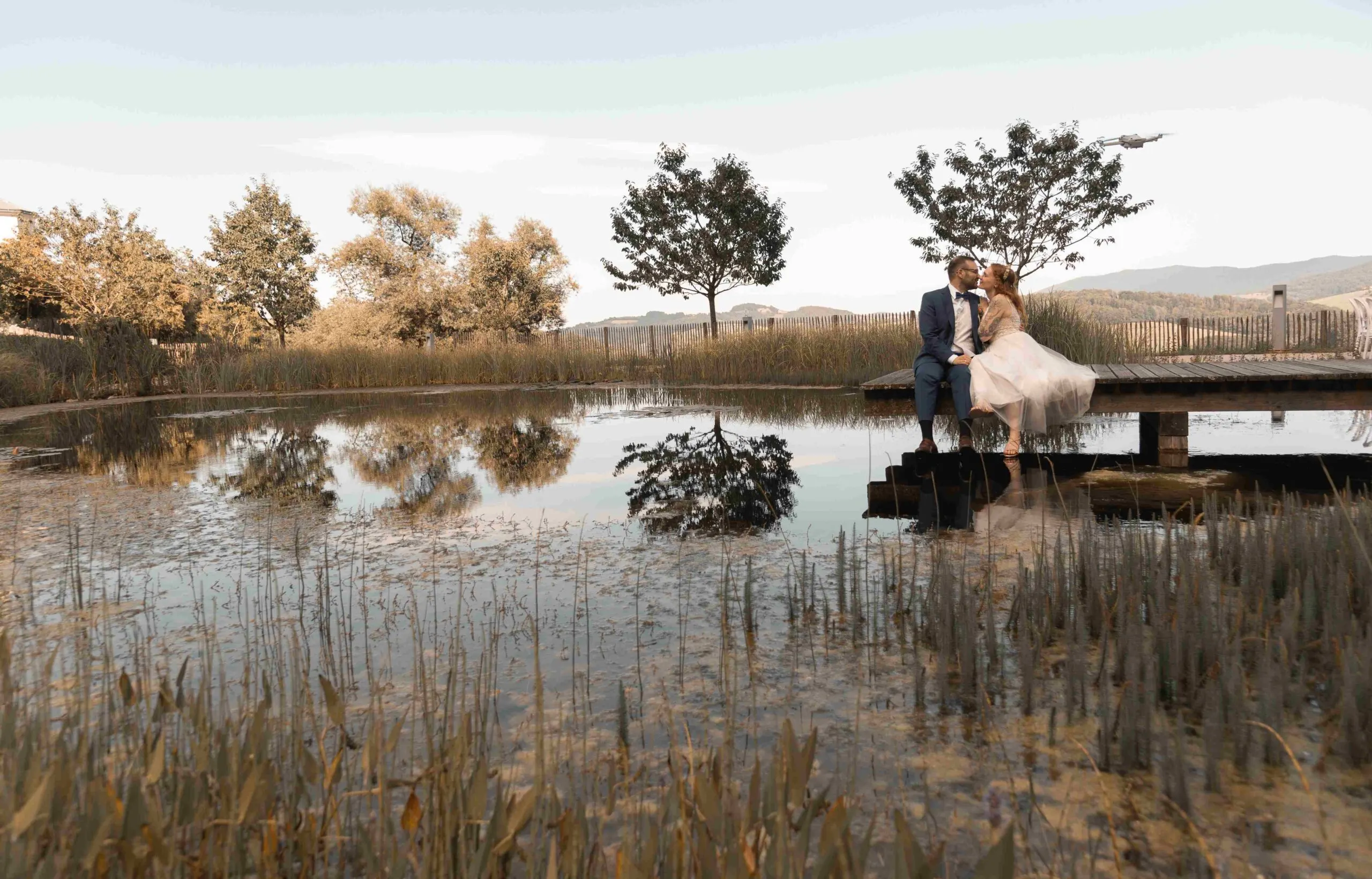 Weite Teichlandschaft: Das Paar sitzt küssend am Holzsteg, Bäume und Himmel spiegeln sich im Wasser.