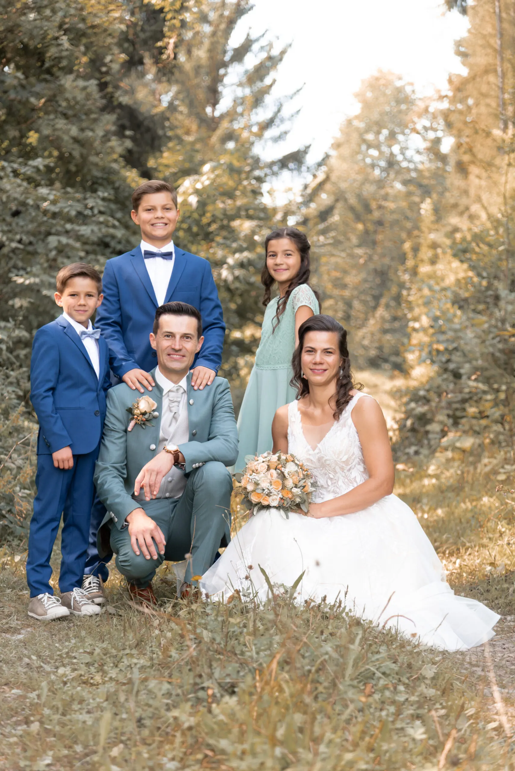 Elisa und Stefan stehen mit ihren drei Kindern Arm in Arm am Waldrand, die Braut hält den apricotfarbenen Strauß