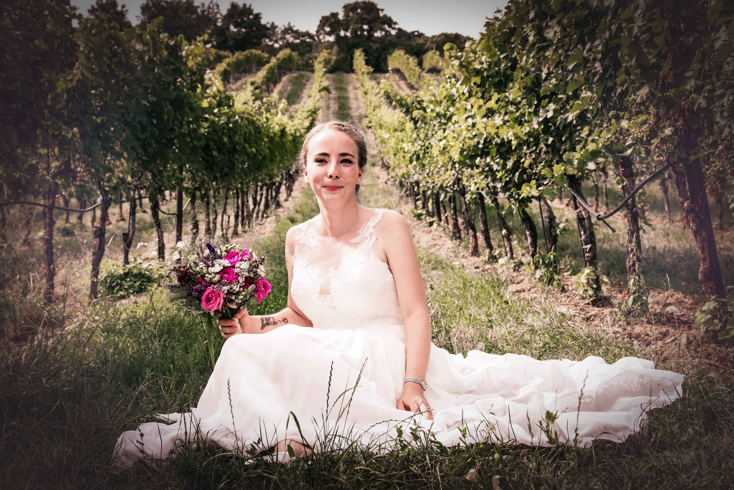 Die Braut sitzt mit pinkem Rosenstrauß im Weingarten