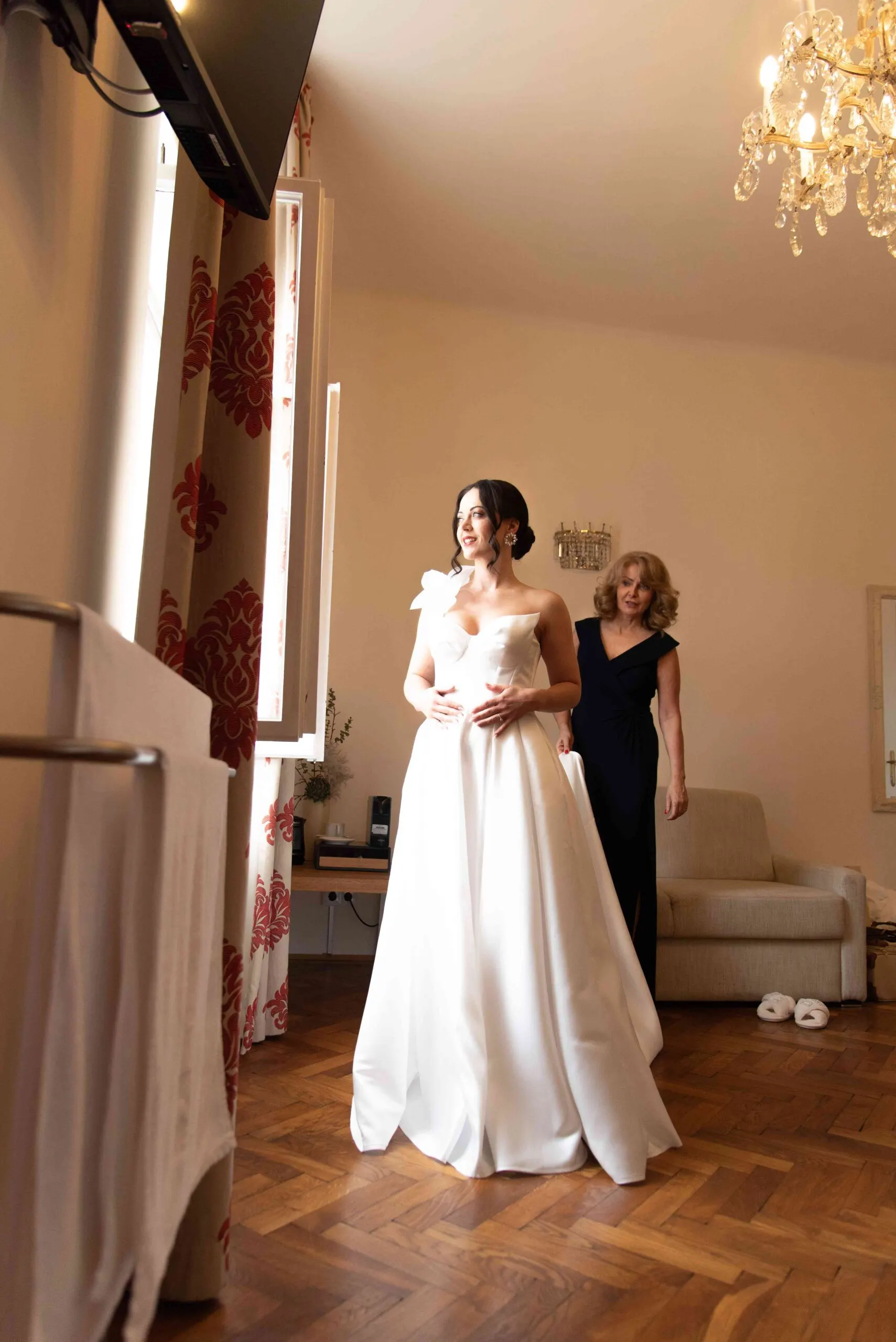Julia im Satinkleid mit Blütenapplikation am Fenster, ihre Mutter hilft im Zimmer mit Kristallluster