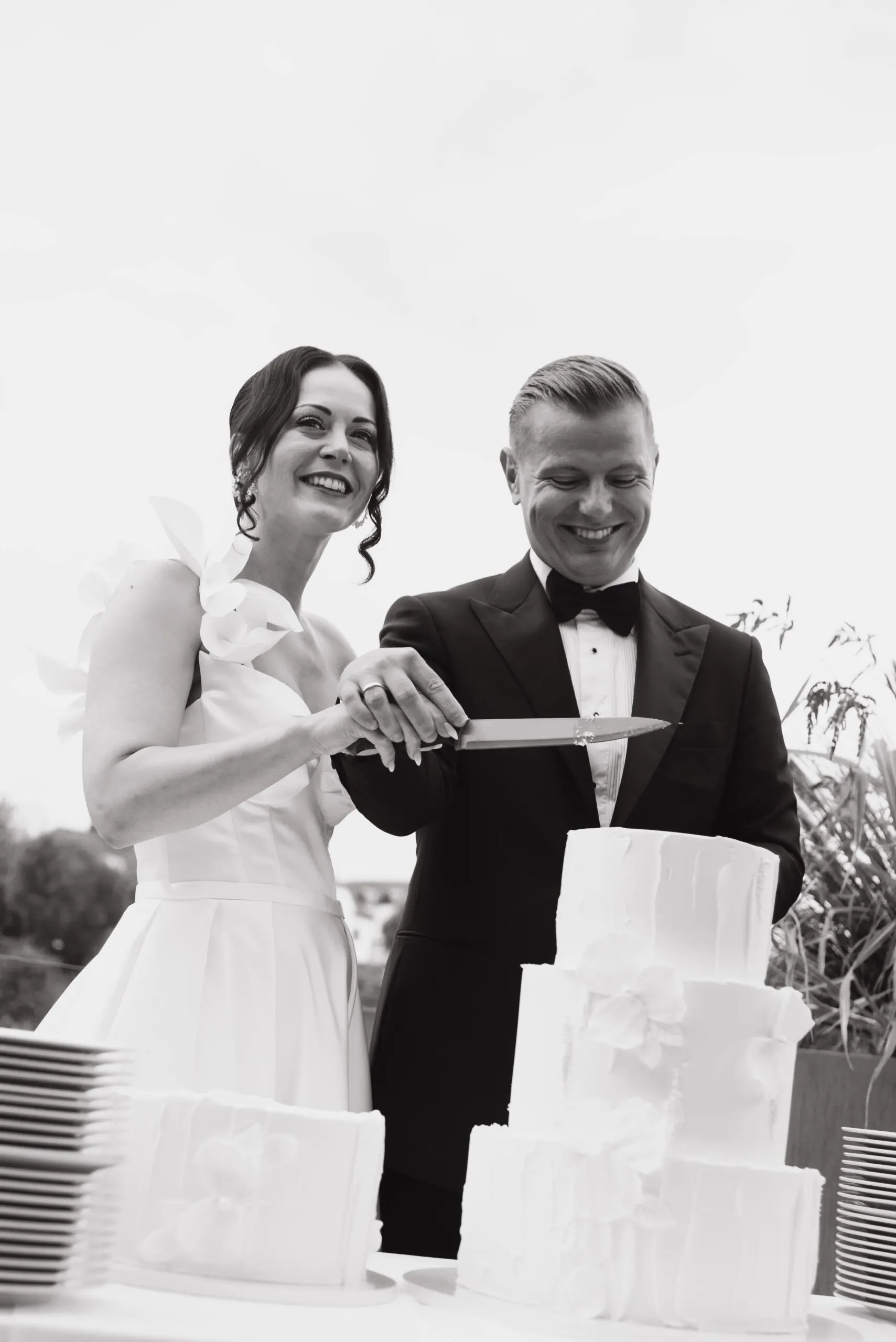 Schwarzweiß: Gemeinsam führen Julia und Denis das Messer zur Torte, dahinter hohe Gräser