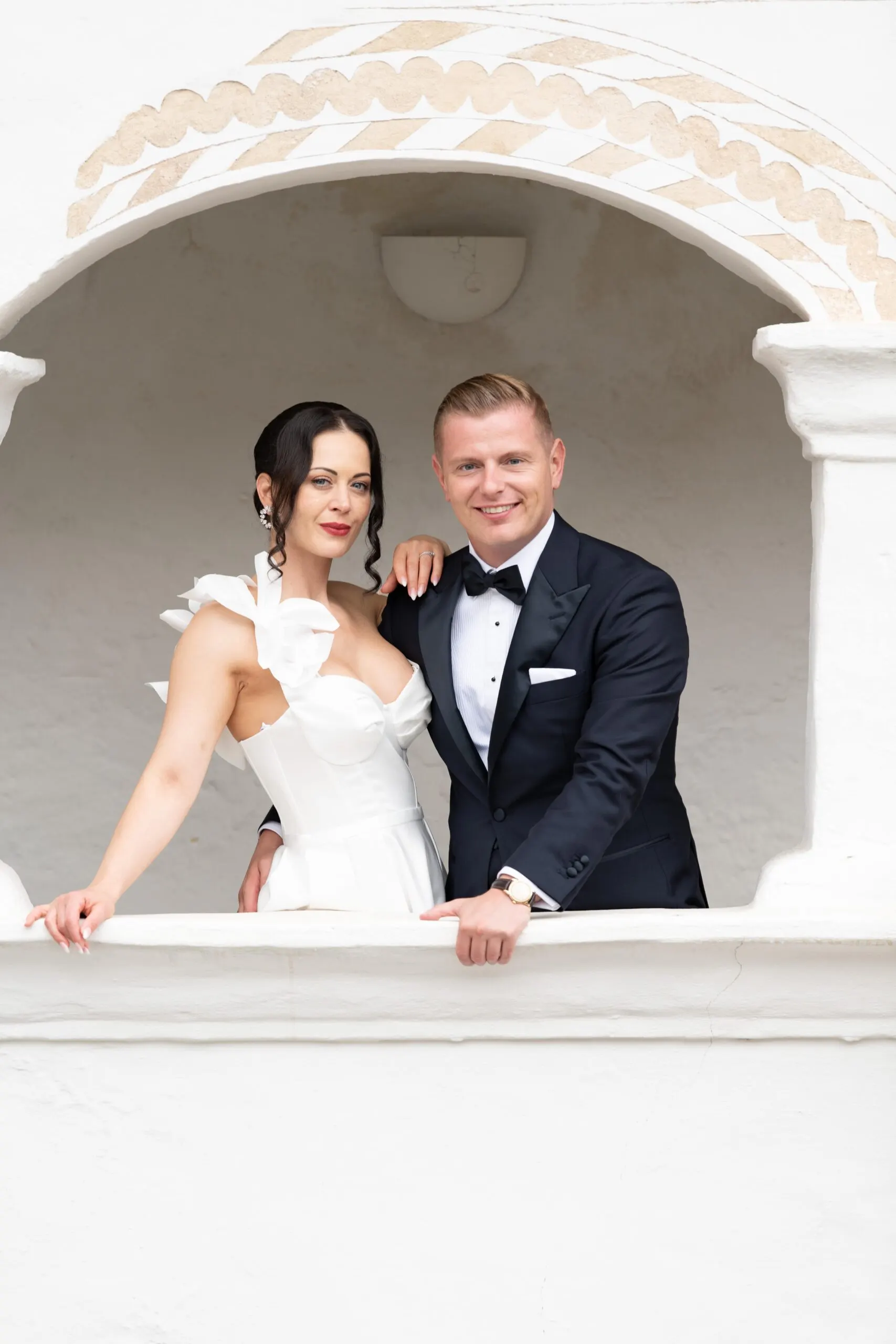 Julia und Denis lächeln aus dem verzierten Bogen der weißen Loggia, ihre Hand auf seiner Schulter