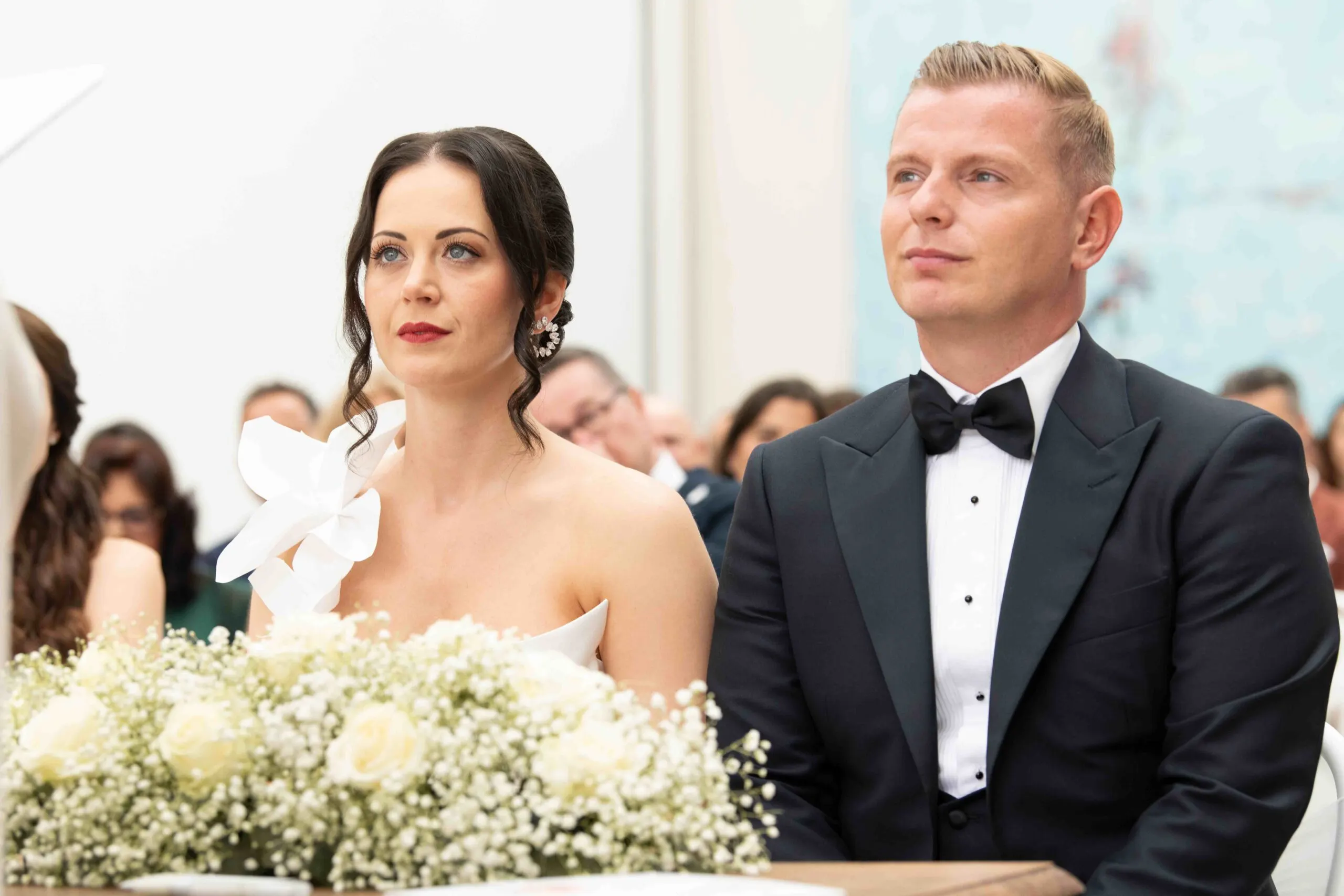 Julia und Denis sitzen bei der Trauung hinter weißen Rosen und Schleierkraut, die Gäste dahinter