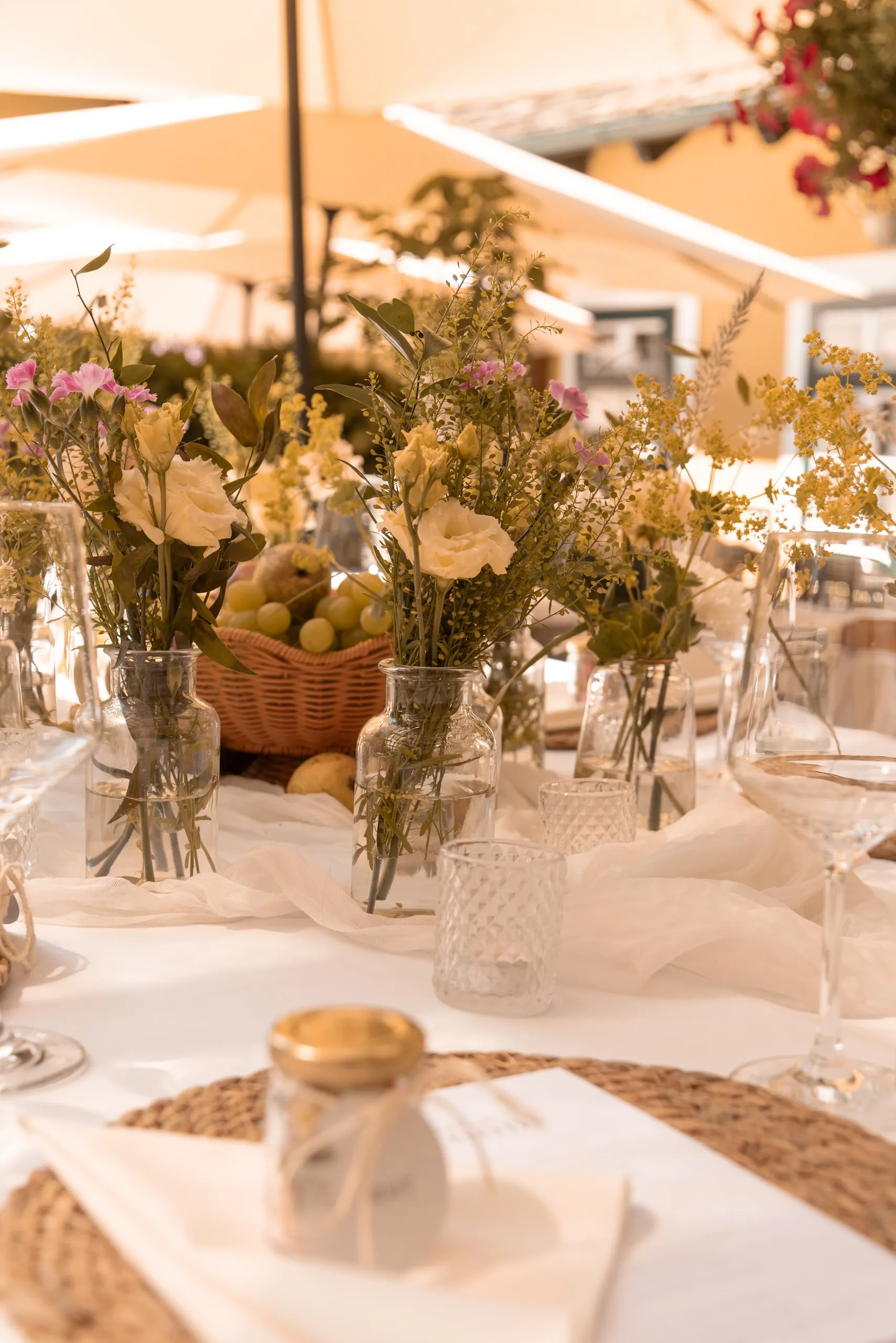 Festlich gedeckte Tafel mit Wildblumen in Glasvasen, einem Obstkorb und Gläsern unter beigen Sonnenschirmen