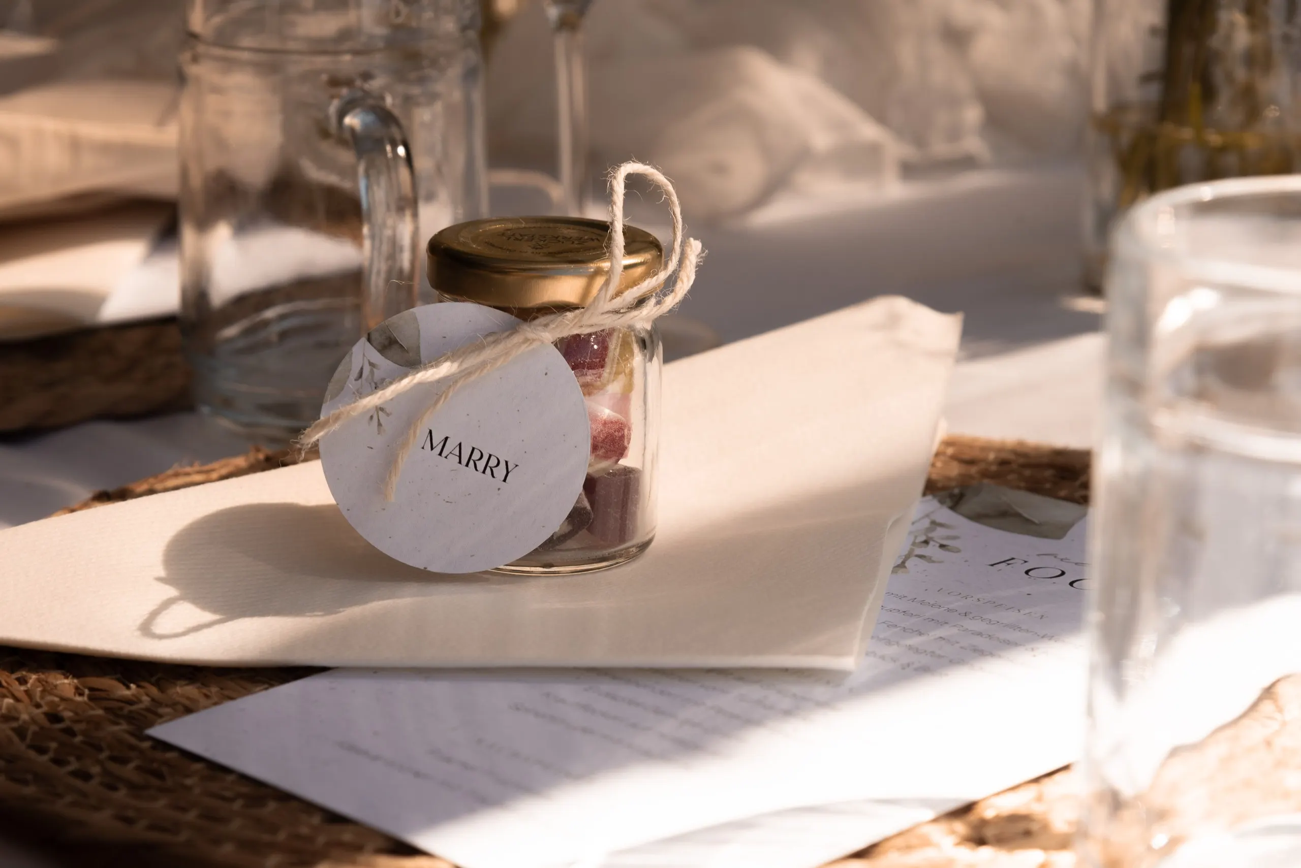 Ein Glas mit dem Anhänger „Marry“ steht auf gefalteter Serviette, Gegenlicht lässt den Hintergrund golden verschwimmen