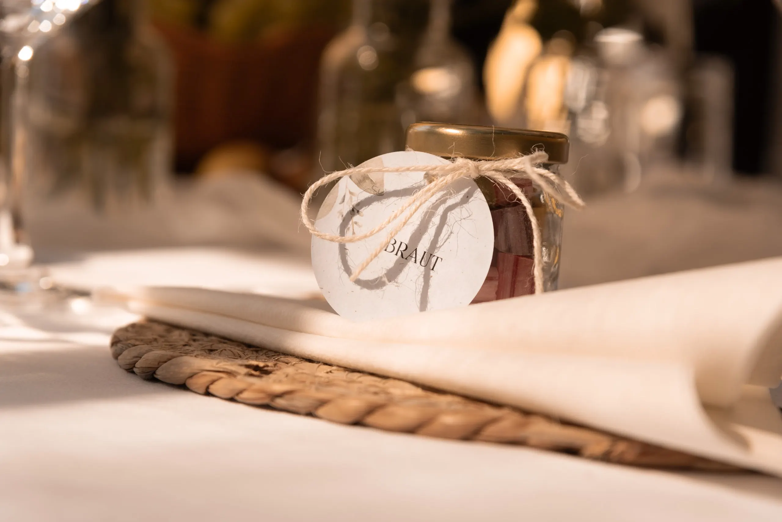 Ein Glas mit dem Anhänger „Braut“ steht auf Serviette und geflochtenem Untersetzer