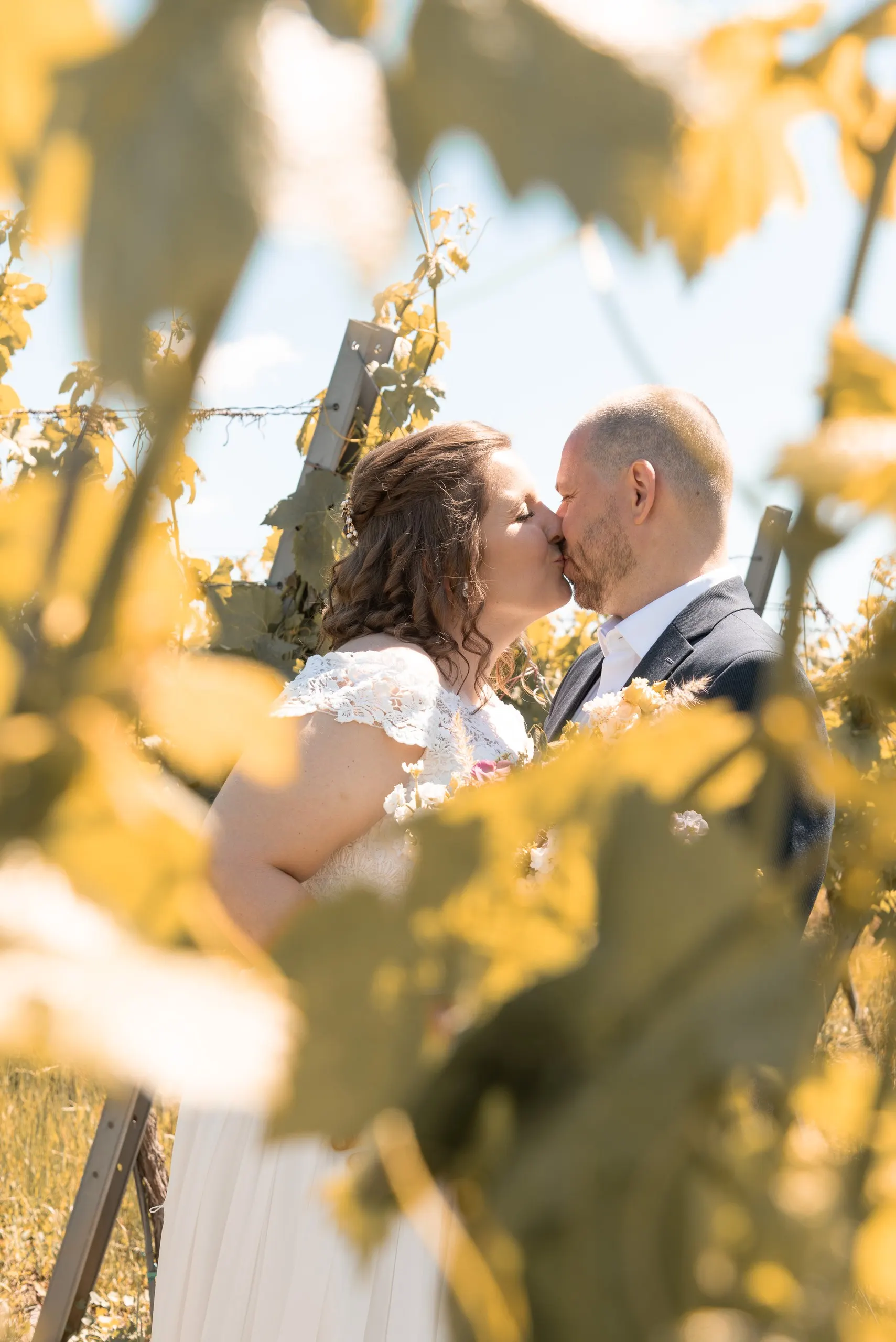 Das Brautpaar küsst sich, durch gelbes Weinlaub hindurch fotografiert