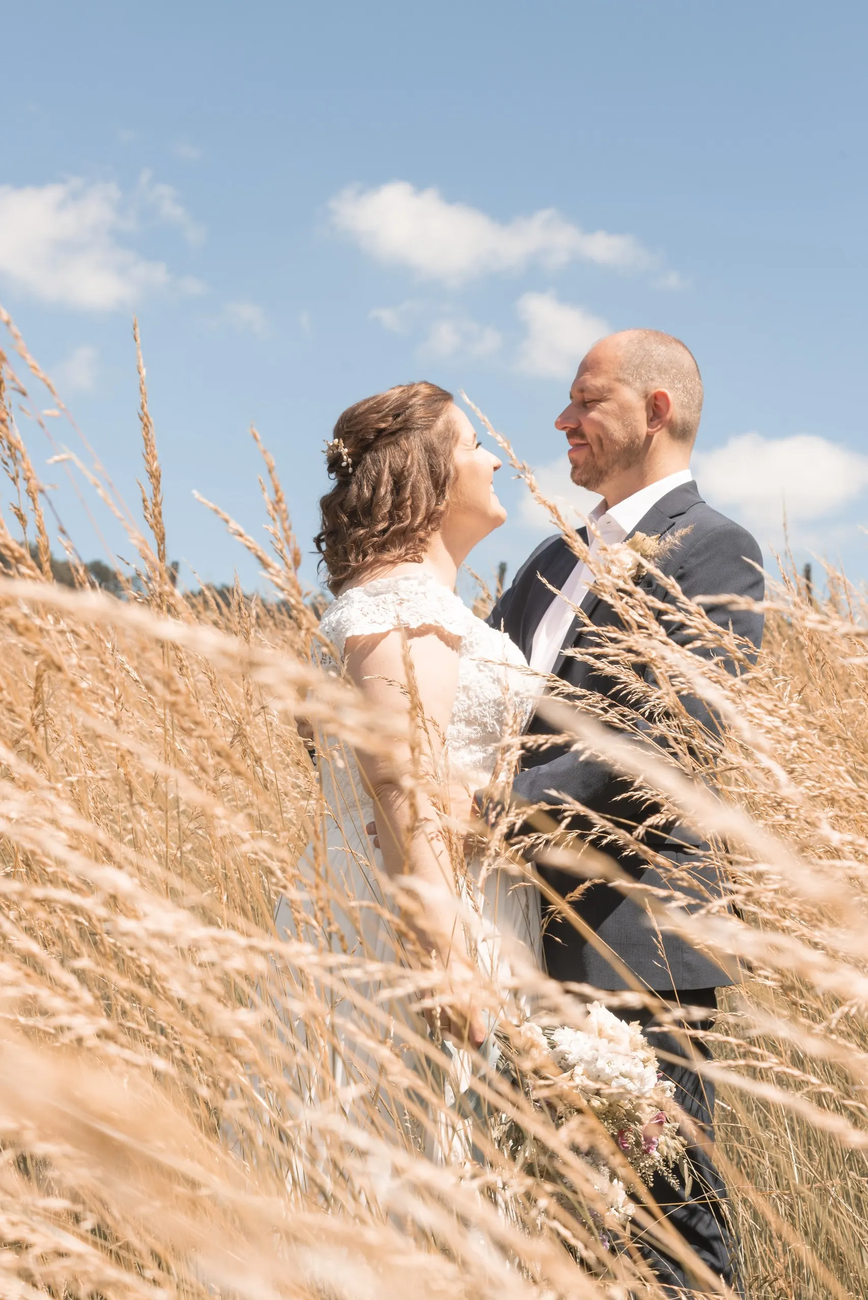 Braut und Bräutigam lachen sich herzlich an, umgeben von hohem goldenem Gras