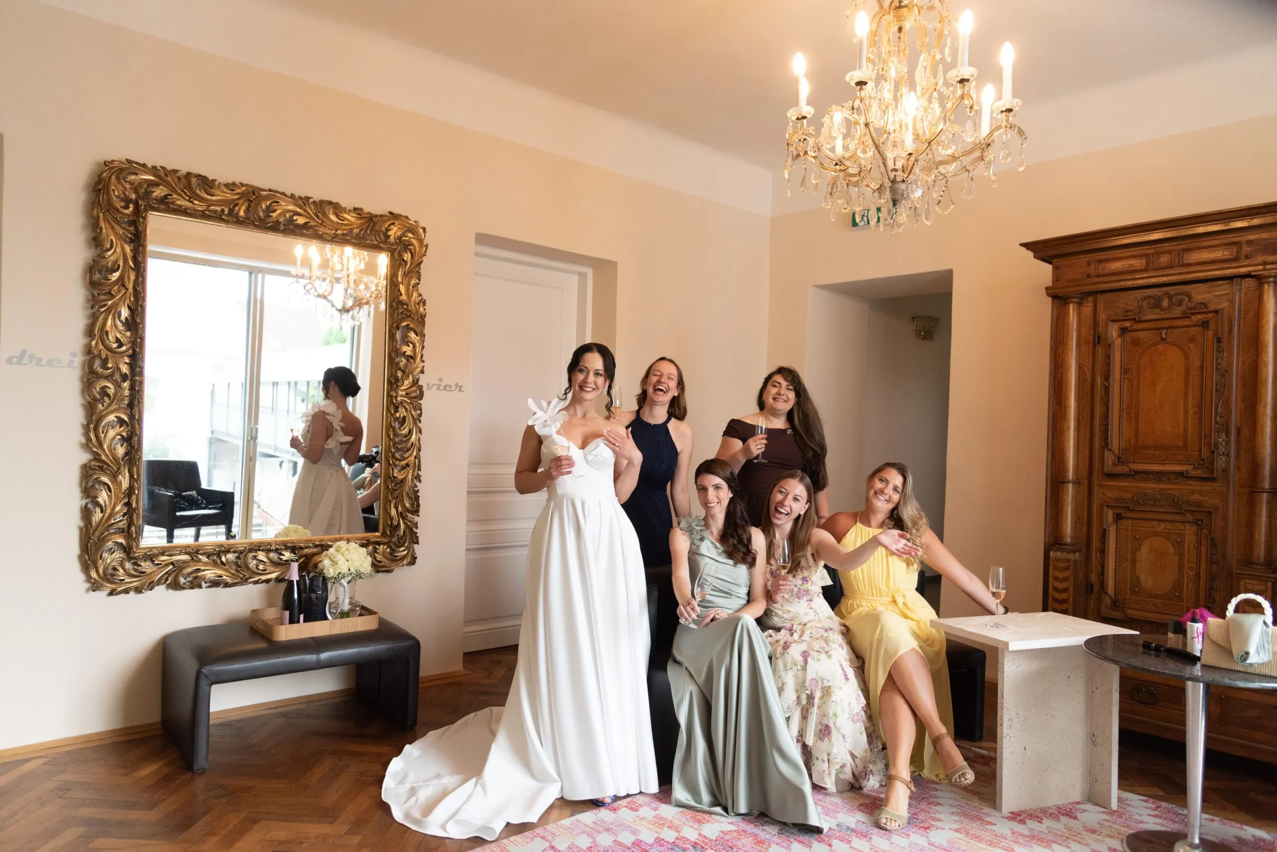 Braut und ihre Freundinnen lachen beim Getting Ready in einem Salon mit Kristallluster