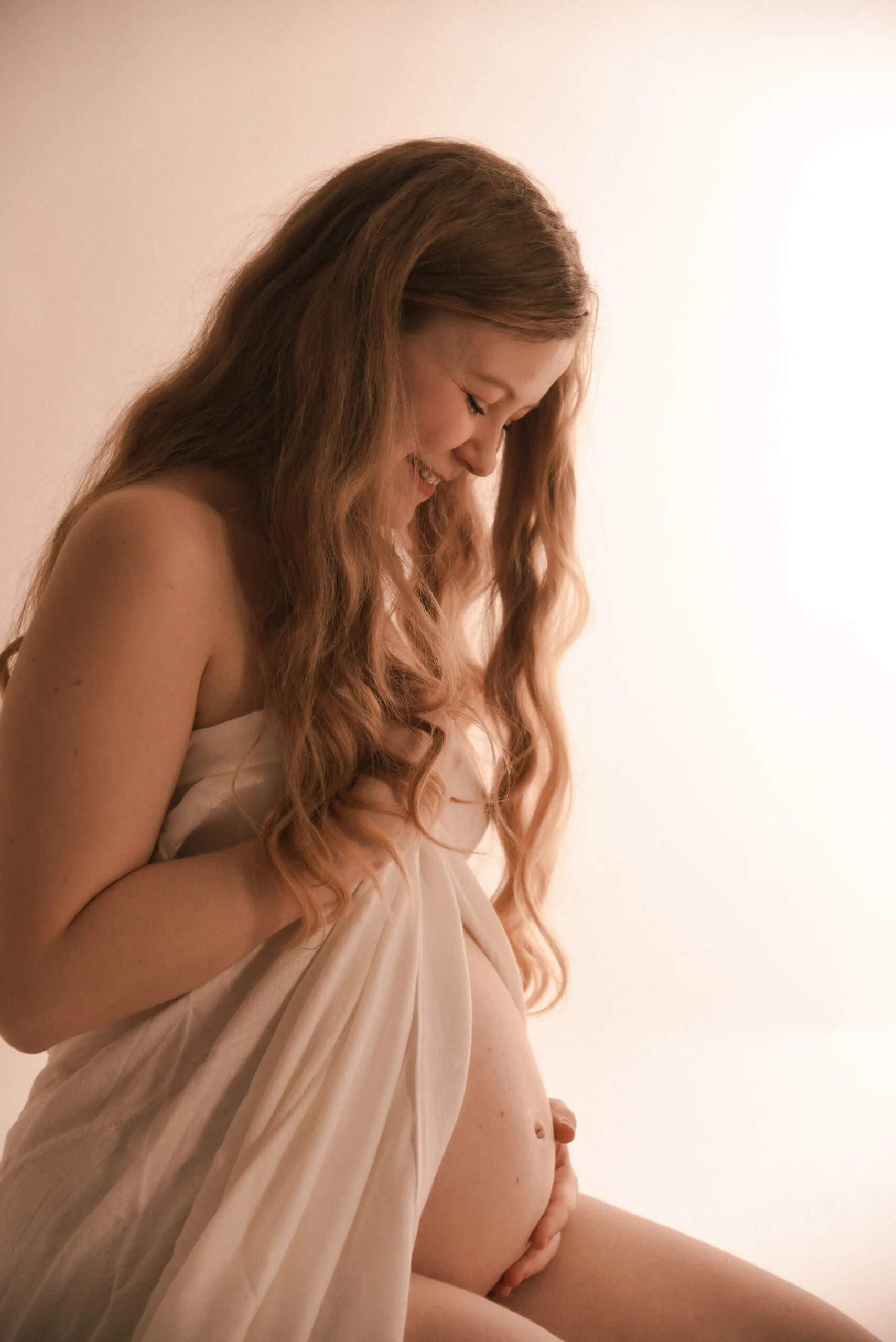 Eine werdende Mutter im Profil, in weißen Stoff gehüllt, blickt lächelnd auf ihren nackten Babybauch.