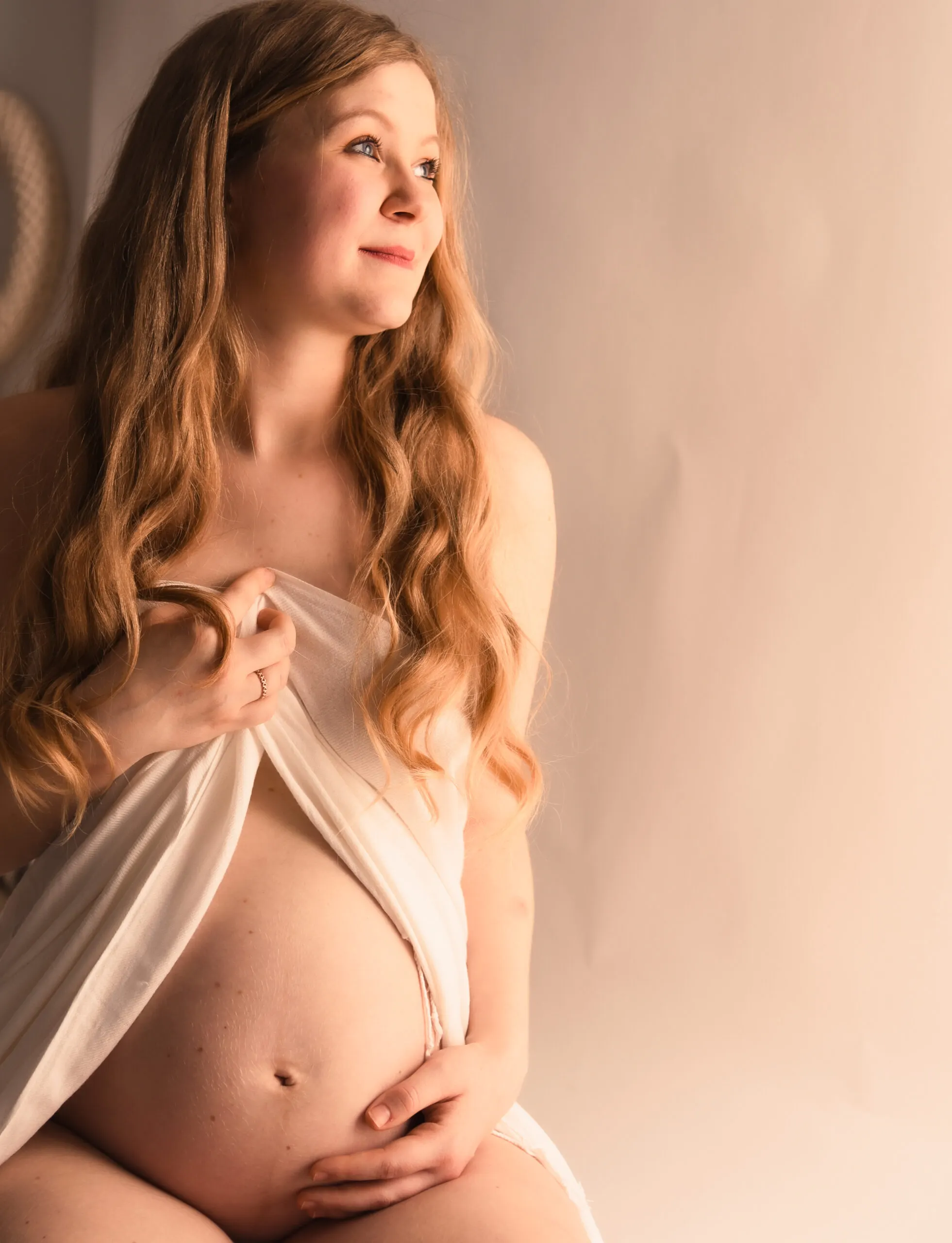 Eine schwangere Frau blickt zum Fensterlicht, ein weißes Tuch vor der Brust, eine Hand am Babybauch.