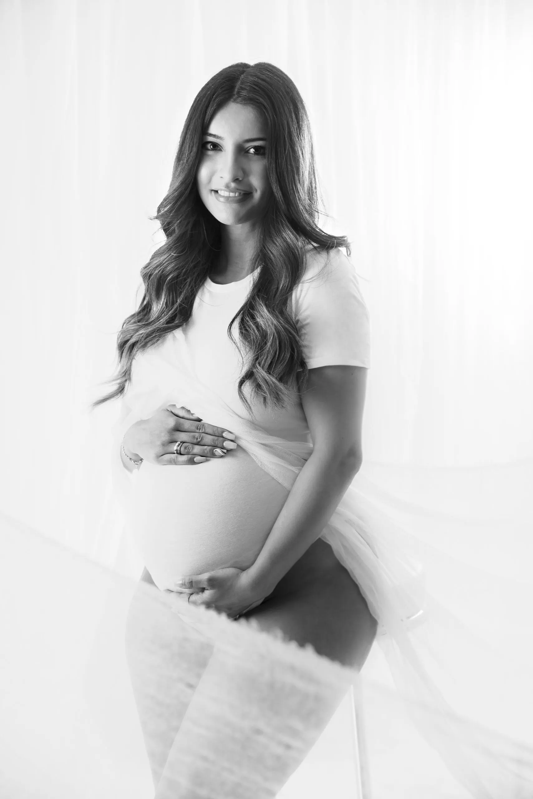 Schwarz-Weiß-Aufnahme: Eine werdende Mutter rafft ihr weißes Shirt über dem Babybauch und lächelt in die Kamera.