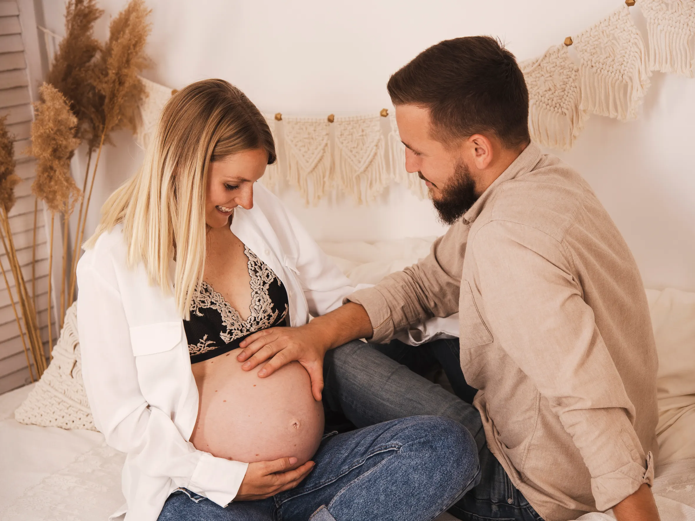 Ein werdender Vater legt die Hand auf den Babybauch seiner lachenden Partnerin, dahinter Makramee-Girlanden.