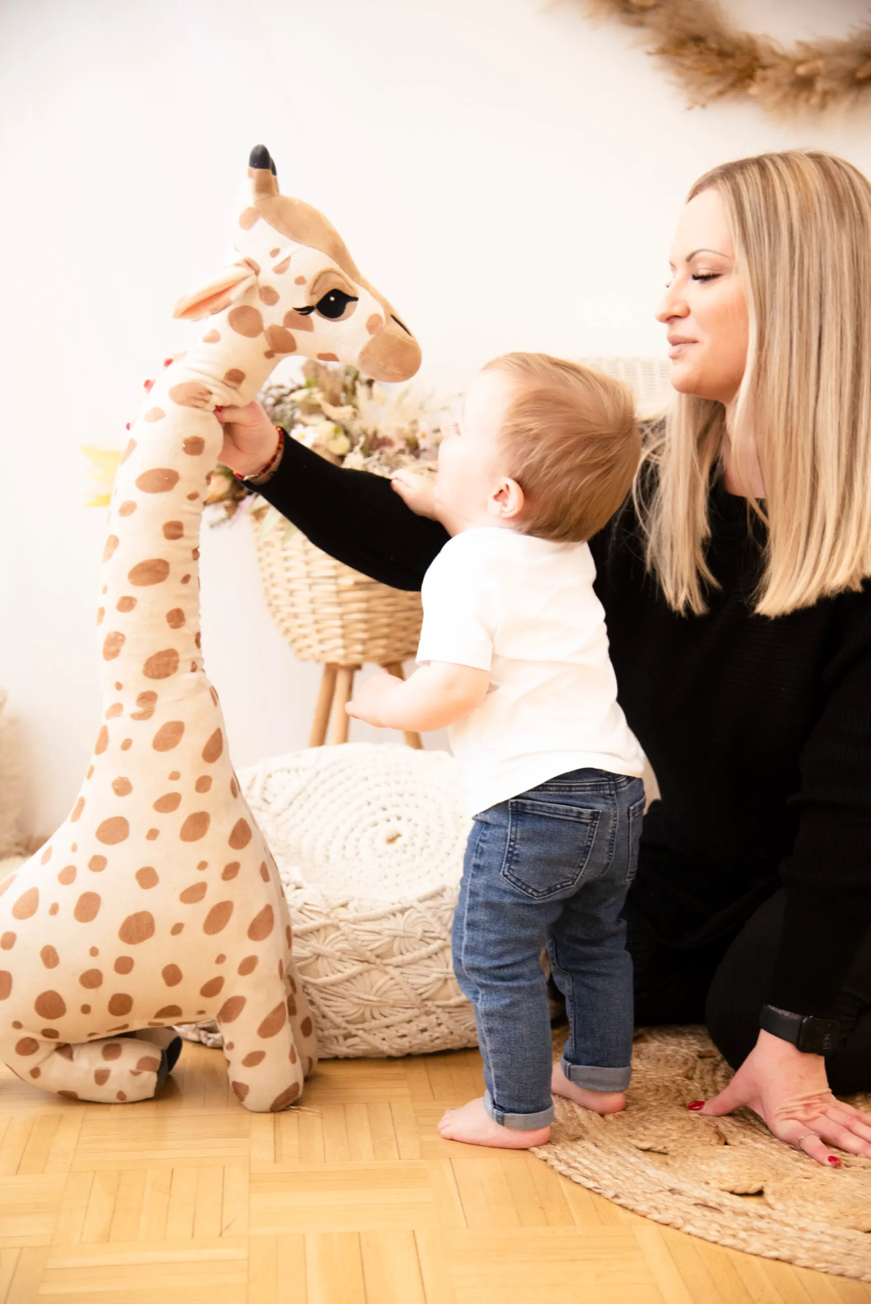 Eine Mutter kniet neben ihrem Kleinkind, das in einem hellen Zimmer eine große Plüschgiraffe umfasst.