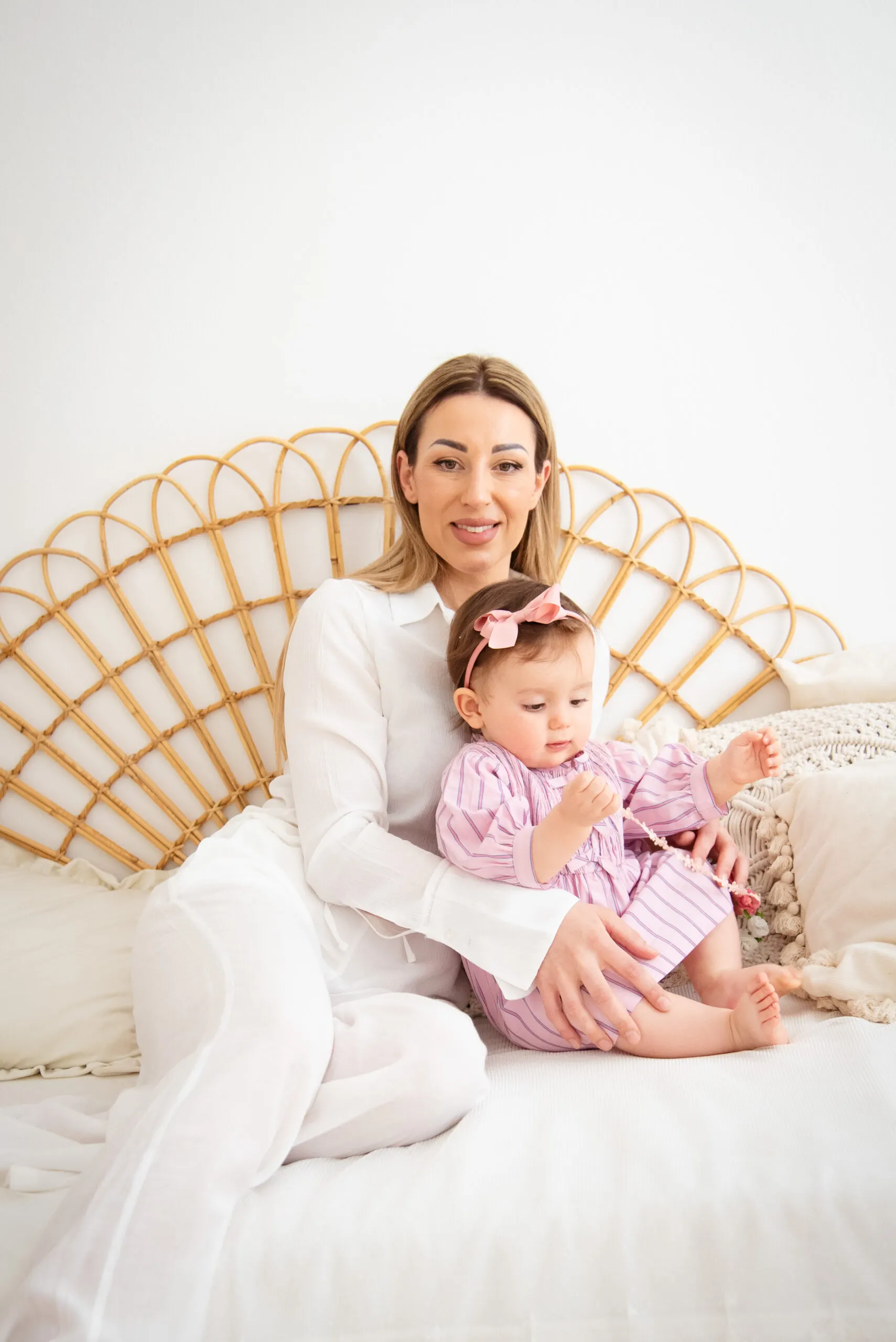 Eine Mutter in Weiß sitzt mit ihrem Baby im rosa gestreiften Kleid vor einem Rattan-Kopfteil.