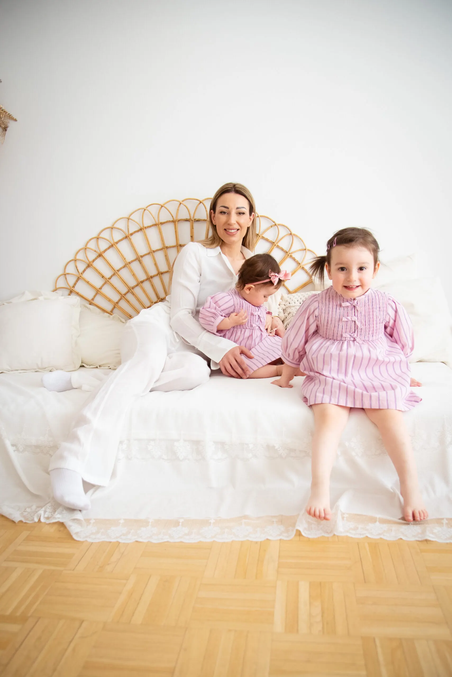 Eine Mutter sitzt mit Baby und lachendem Kleinkind in gleichen rosa Kleidchen auf einem hellen Bett.