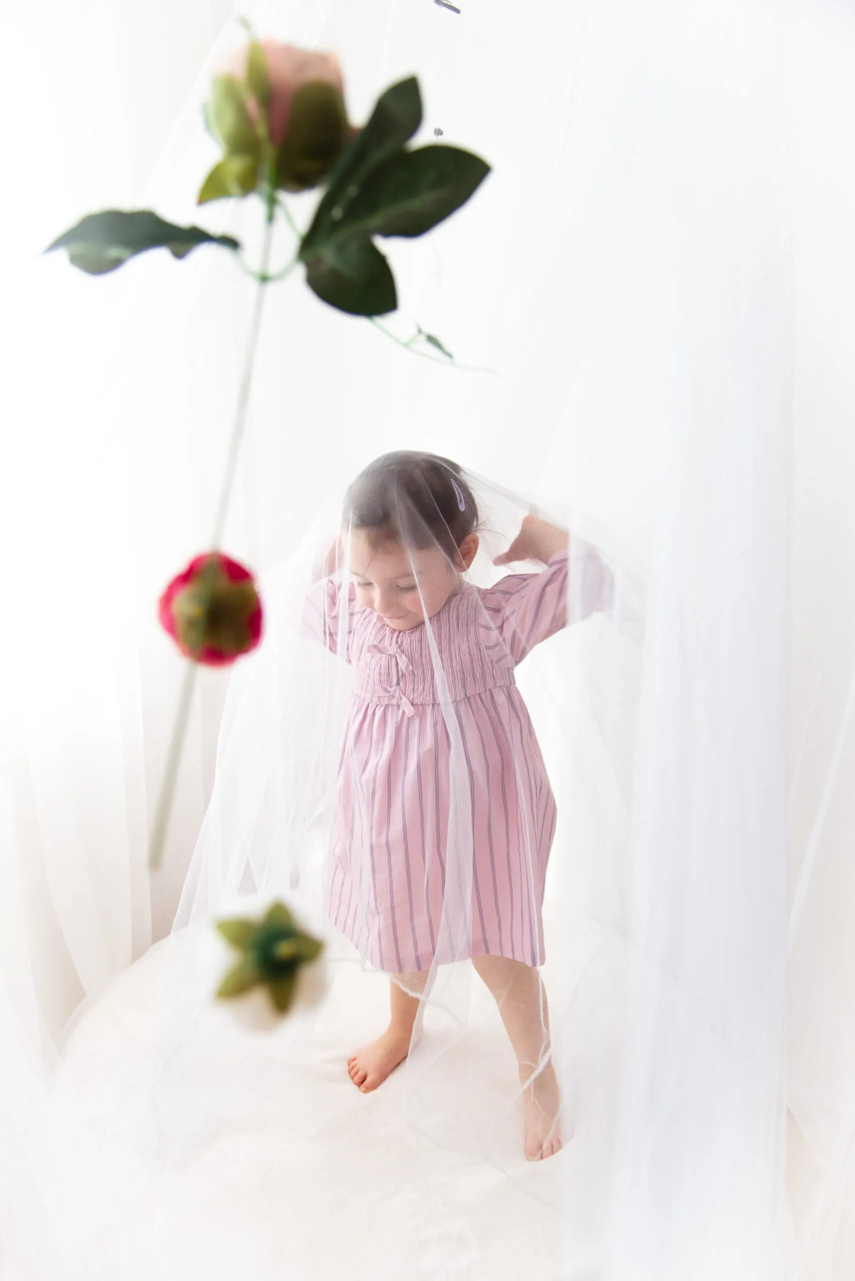 Ein Kleinkind im rosa Kleid zieht sich einen zarten Tüllvorhang über den Kopf, davor hängende Blüten.