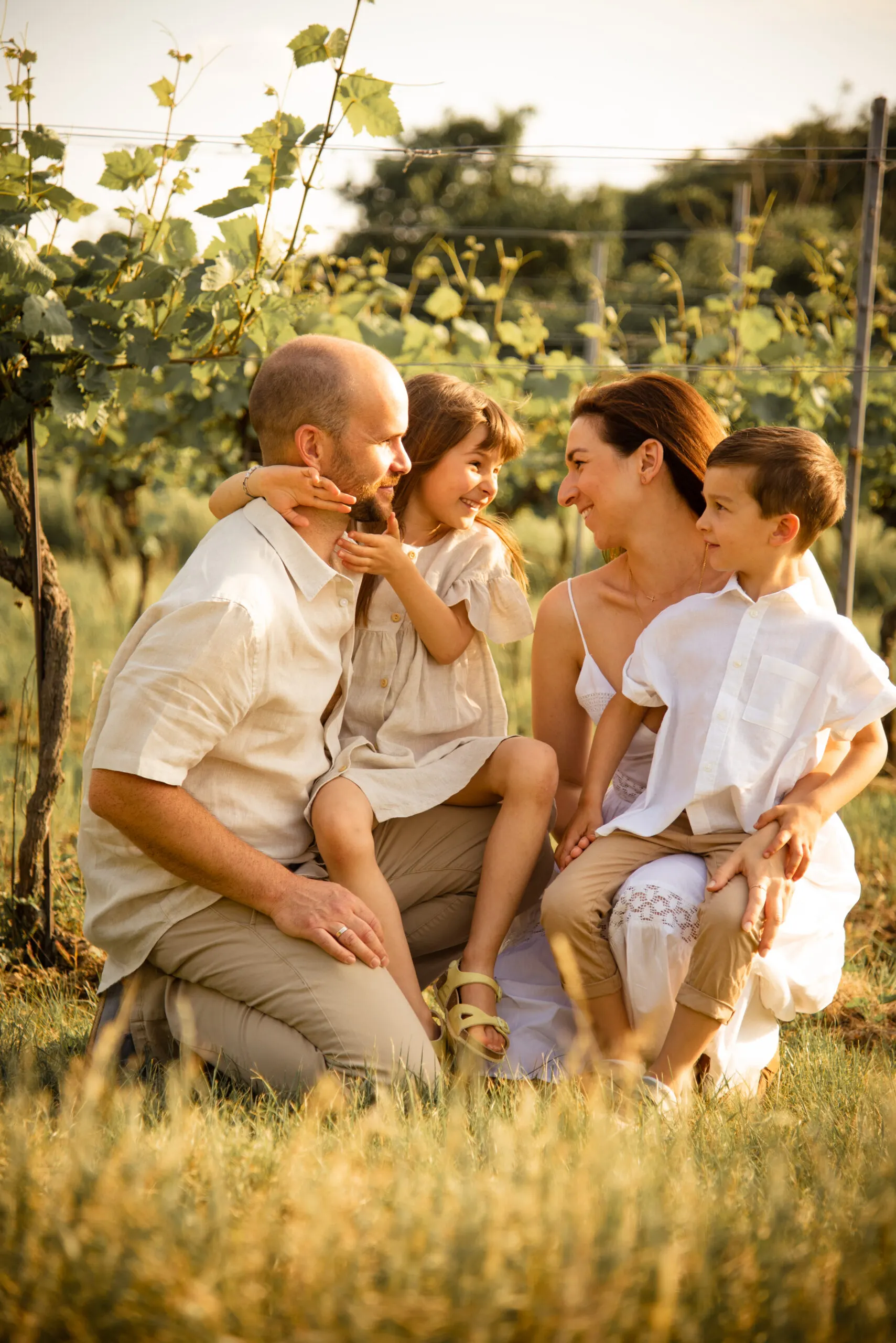 Eine Familie kniet im goldenen Licht eng beisammen zwischen Rebzeilen und lacht einander an.