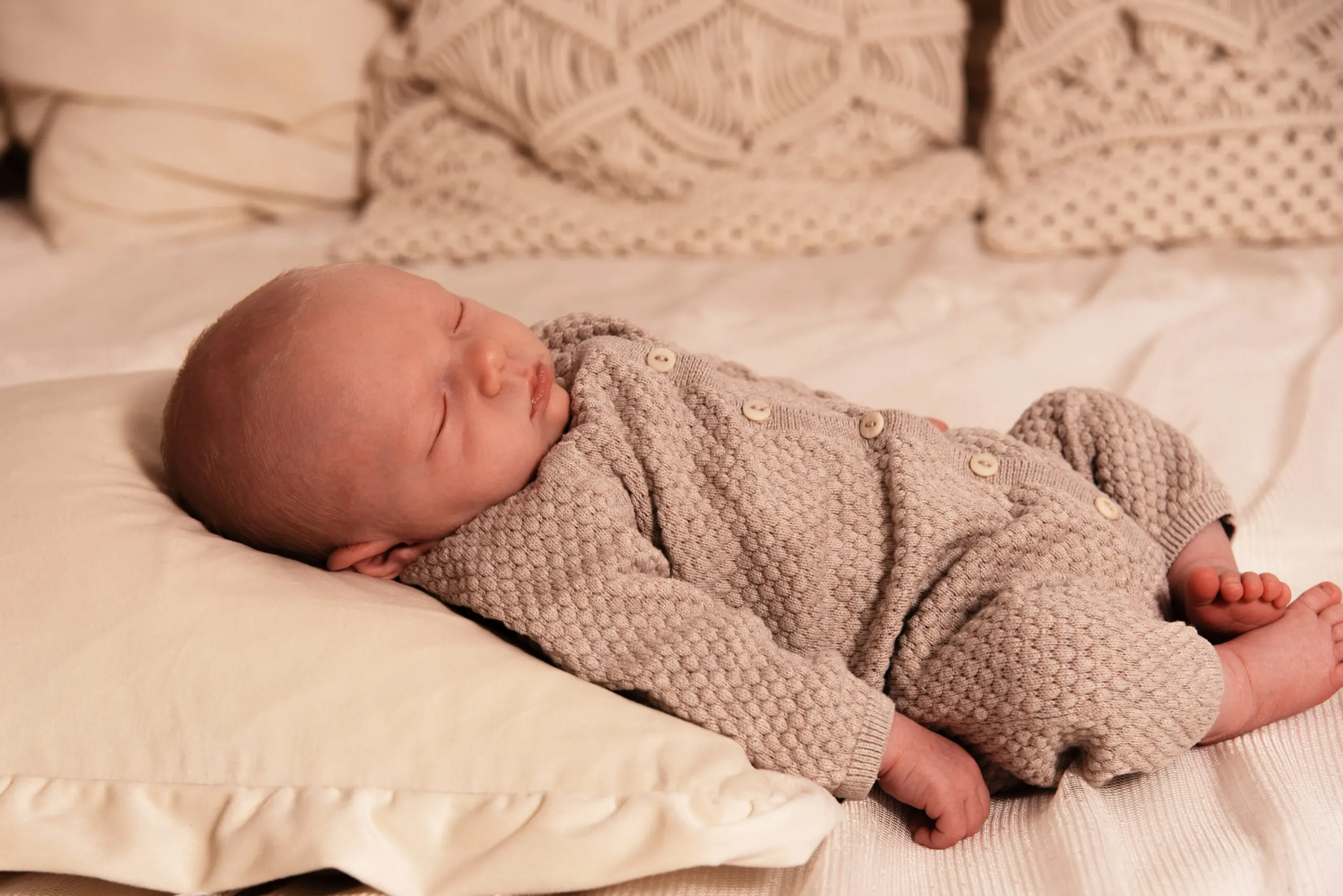 Neugeborenes schläft im beigen Strickoverall auf einem cremefarbenen Polster vor Makramee-Kissen.