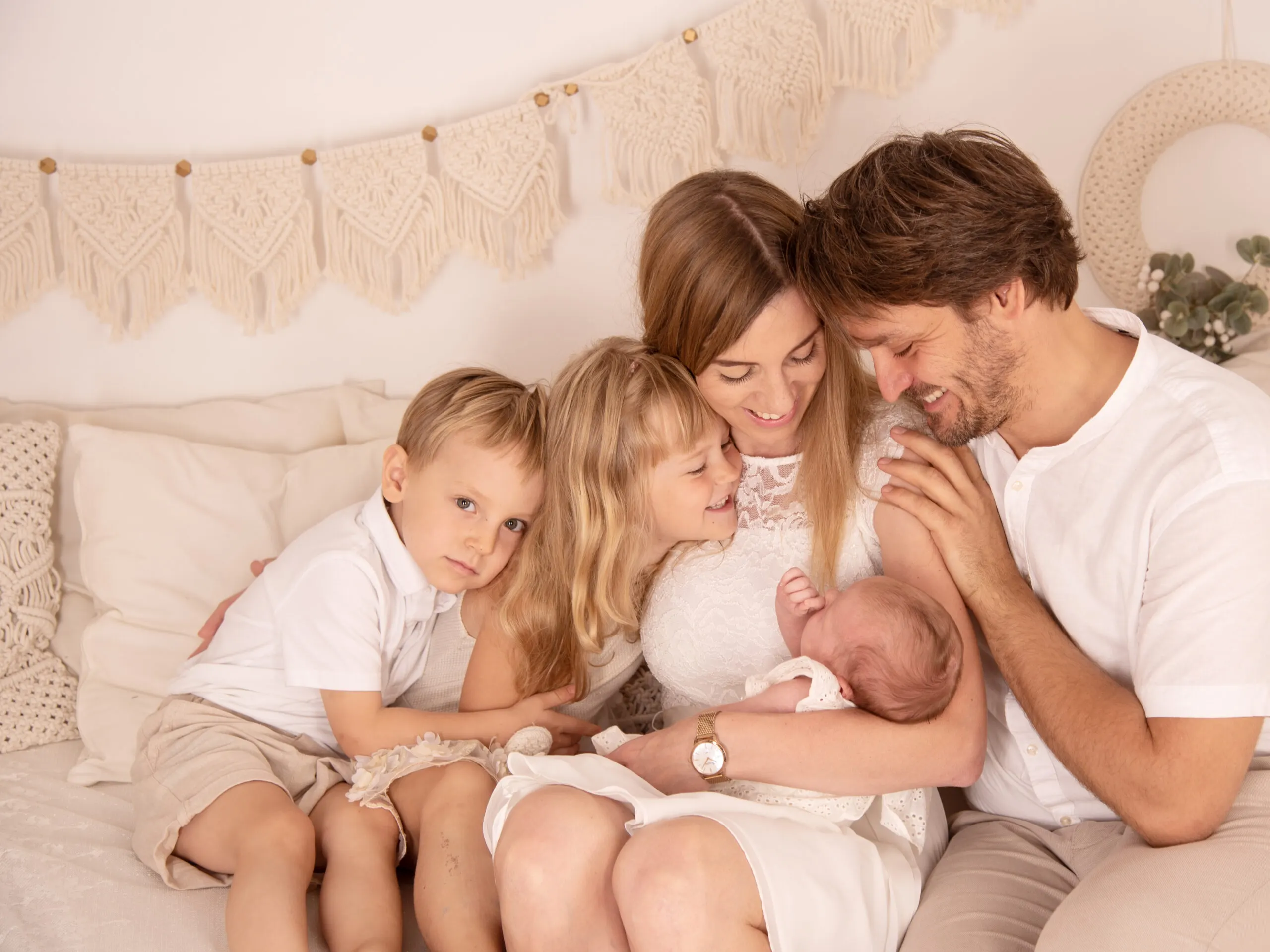 Eltern mit drei Kindern kuscheln in hellen Outfits auf dem Bett, die Mutter hält das Neugeborene.