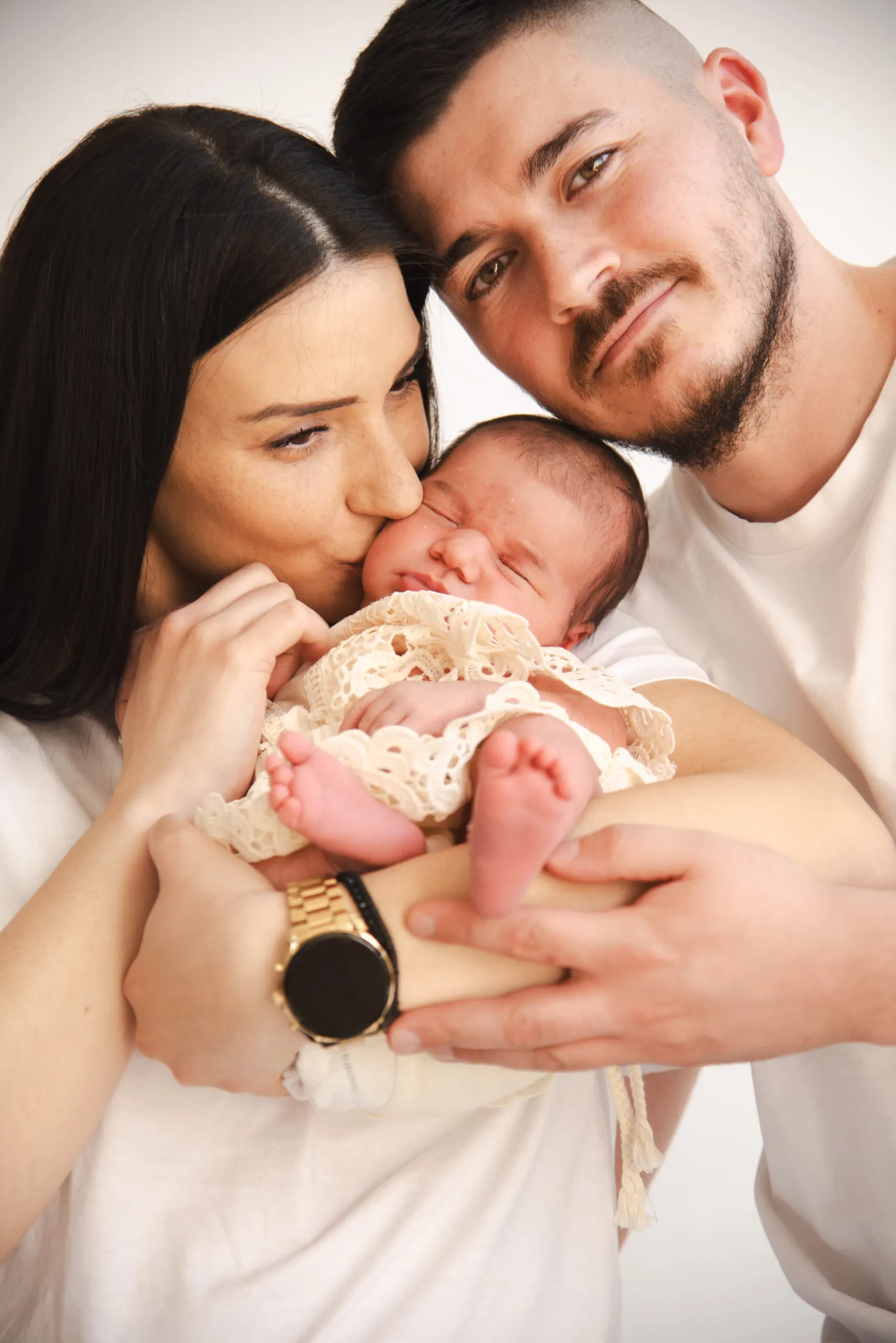 Mutter küsst ihr Neugeborenes im Spitzenstrampler auf die Wange, der Vater blickt in die Kamera.