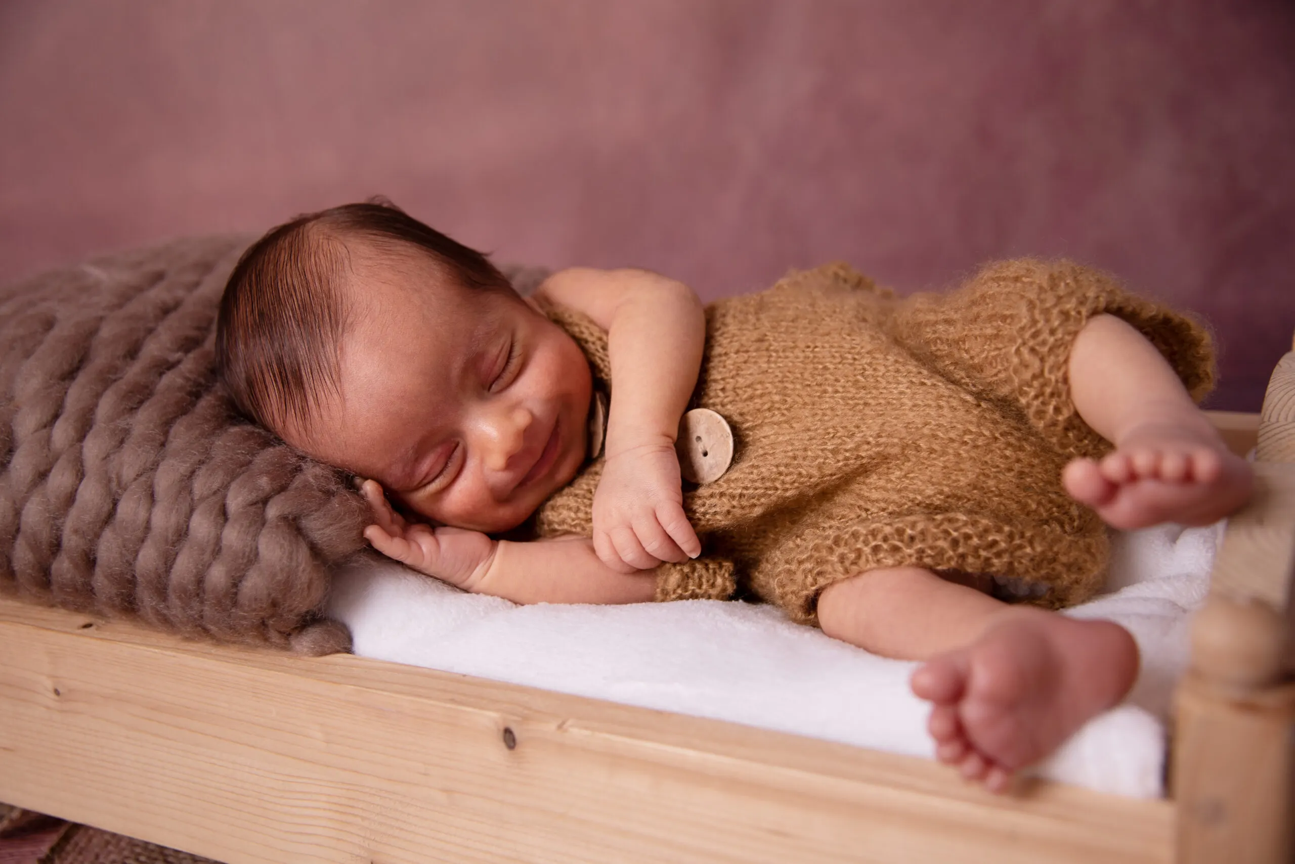 Lächelnd schlafendes Neugeborenes im braunen Strickromper liegt seitlich in einem kleinen Holzbettchen.